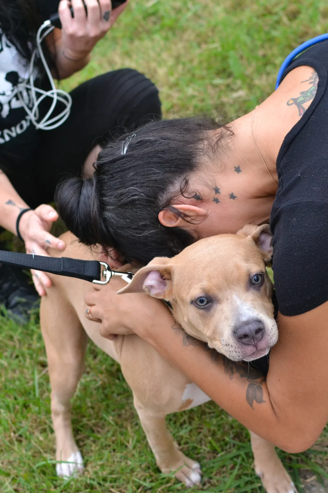 Person hugging a tan and white dog outdoors on grass, with another person holding a leash nearby.