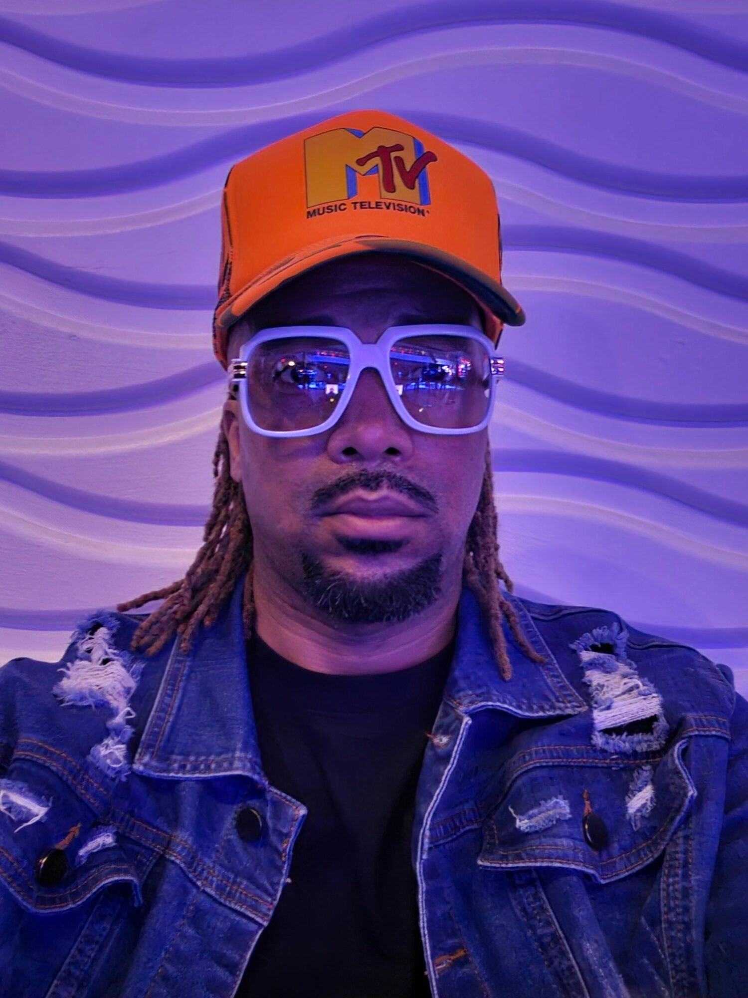 Person wearing glasses and an orange cap with a logo, in front of a purple wavy background.
