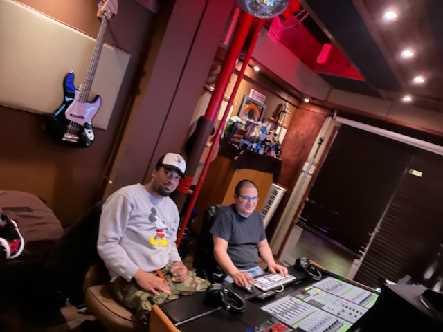 Two men sit at a mixing console in a recording studio, with guitars hanging on the wall behind them.