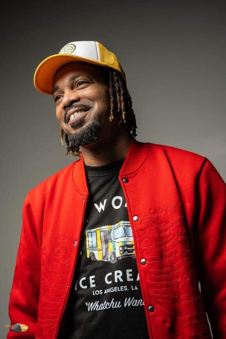 Man wearing a yellow and white cap, red jacket, and black shirt, smiling against a gray background.