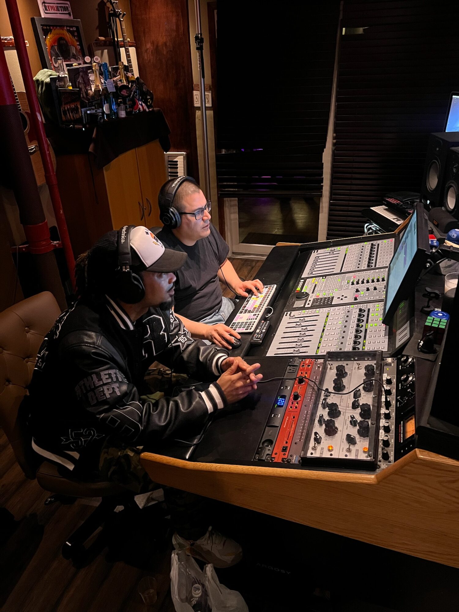 Two people in a recording studio working with audio equipment, one wearing headphones and a cap, the other with glasses, at a mixing console.