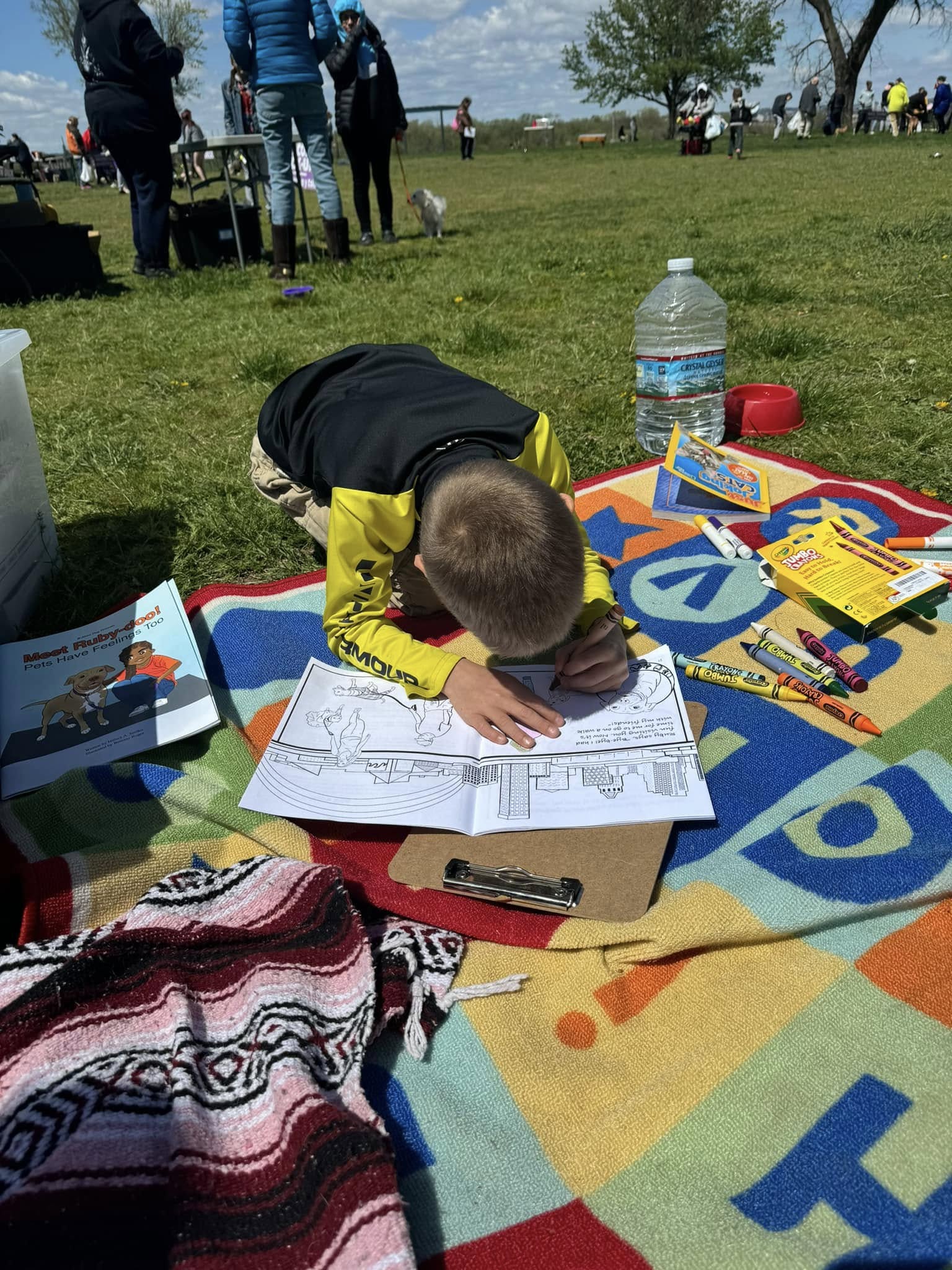 Child lying on a colorful blanket outdoors, drawing on paper with markers, surrounded by water bottles and books, with people in the background.