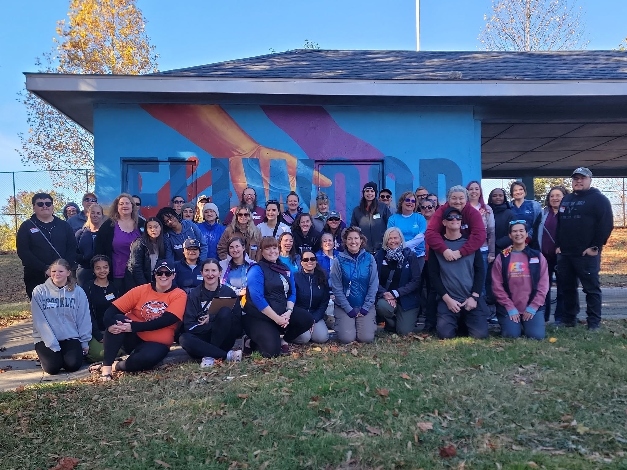 Group of people standing outdoors in front of a colorful mural on a building, with trees and grass nearby.