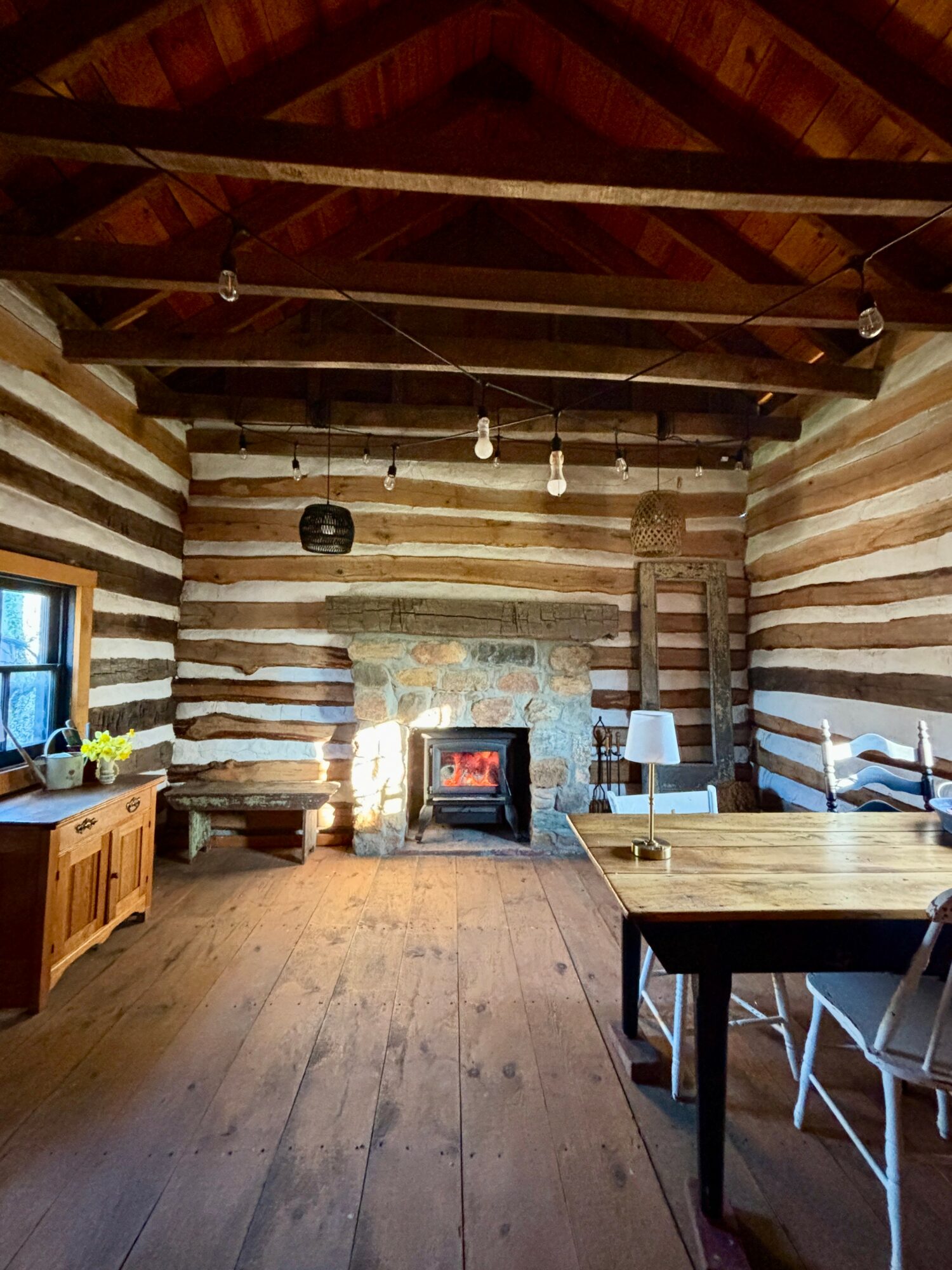 Interior of a log cabin with wooden walls, ceiling, and floor, fireplace, and furniture including a table and chairs.