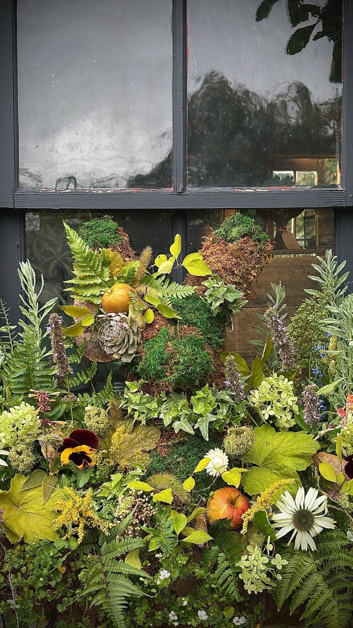 Window with a colorful flower arrangement including yellow, white, and purple flowers, and green foliage, seen through glass panes.