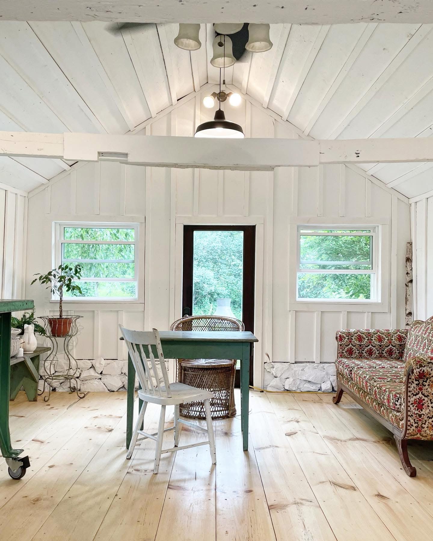 Room with white wooden walls, two windows, a door, a table with chairs, and a patterned sofa, ceiling light fixture.