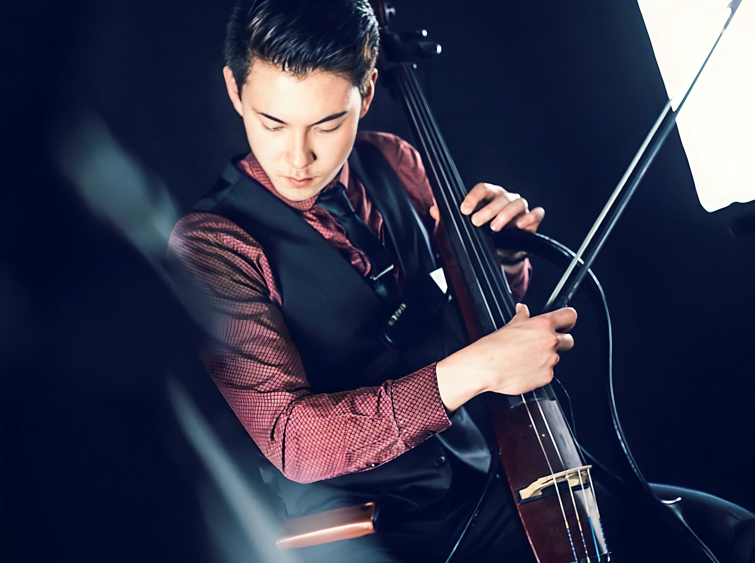 Person playing a cello, focused on the instrument, with a dark background and bright light source, wearing a dark vest and red shirt.