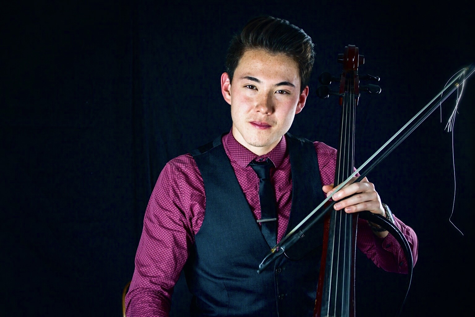Young person holding a violin and bow, wearing a purple shirt and dark vest, against a dark background.