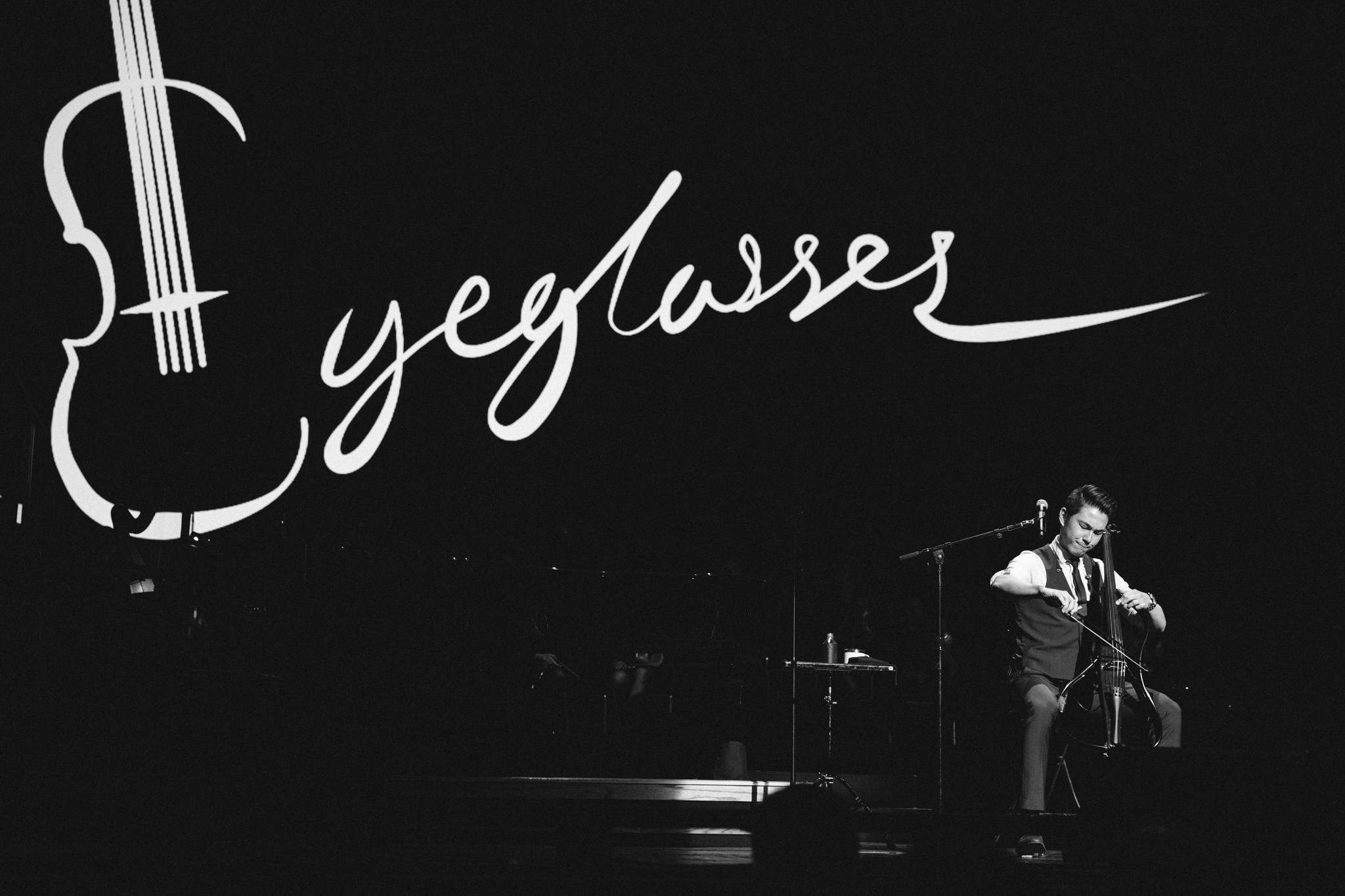 A person playing a cello on stage with a large violin outline and the word 'eylasses' in cursive, black background.