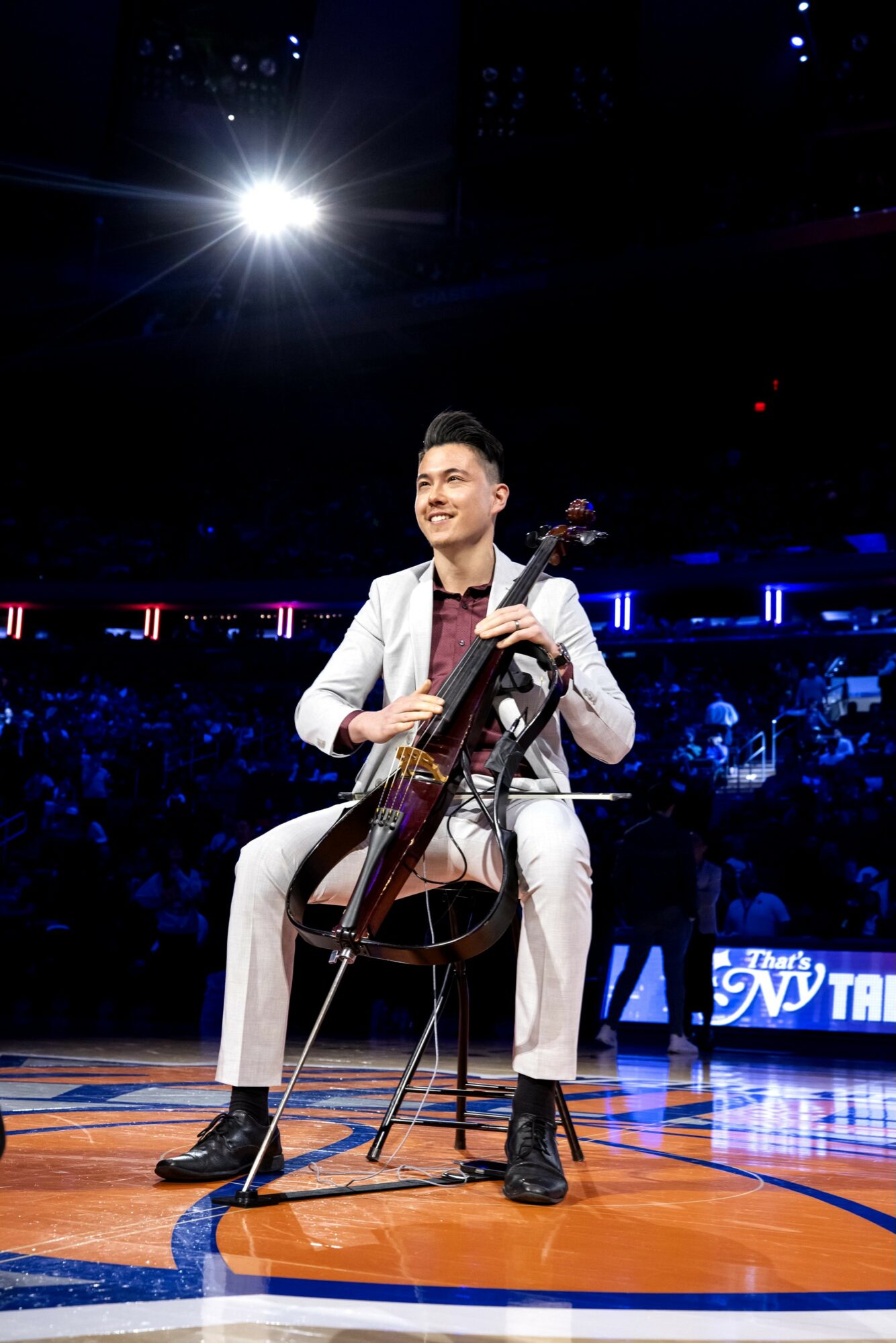 A man in a white suit playing a cello on stage with bright lights and an audience in the background.