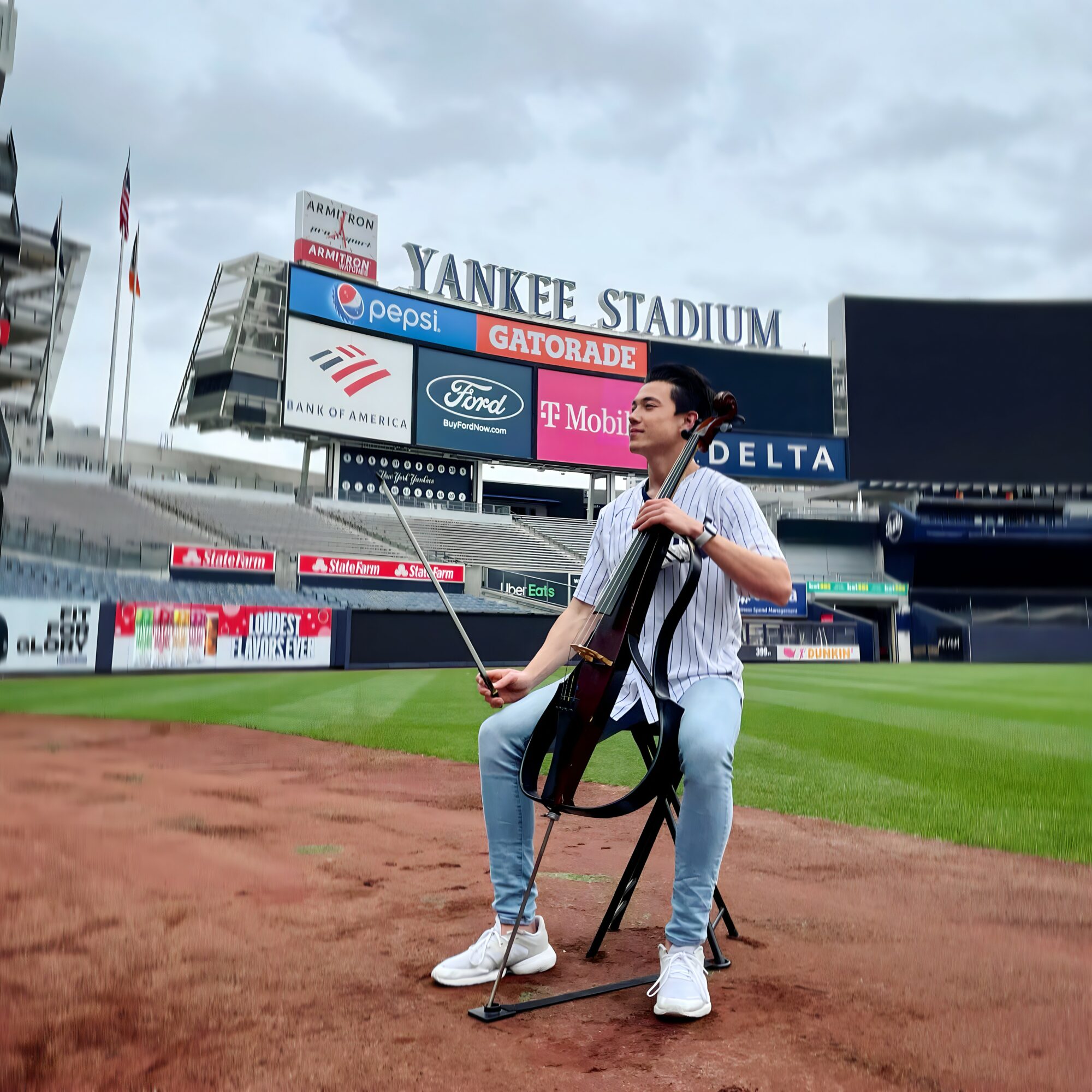 Person sitting on a chair on baseball field with a cello, at Yankee Stadium, cloudy sky, stadium signs visible.