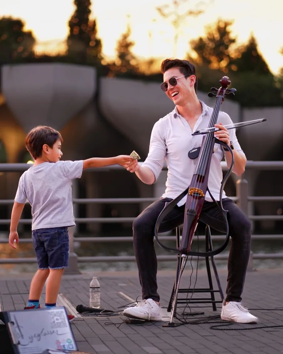 A man playing a cello and a young boy exchanging a handshake outdoors during sunset, with trees and a fence in the background.