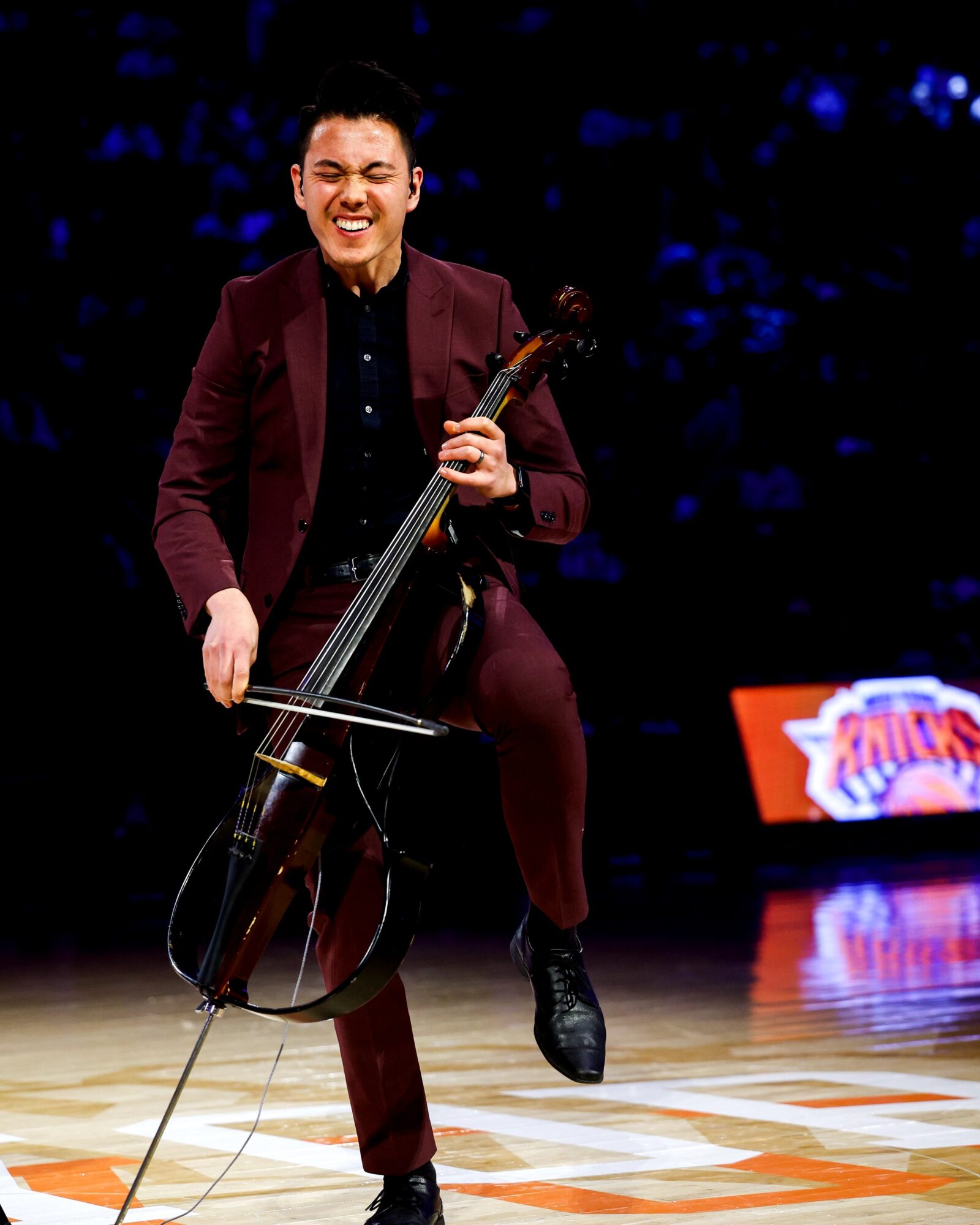 Person playing a cello on stage, smiling, wearing a dark suit, with a dark background and a bright sign in the background.