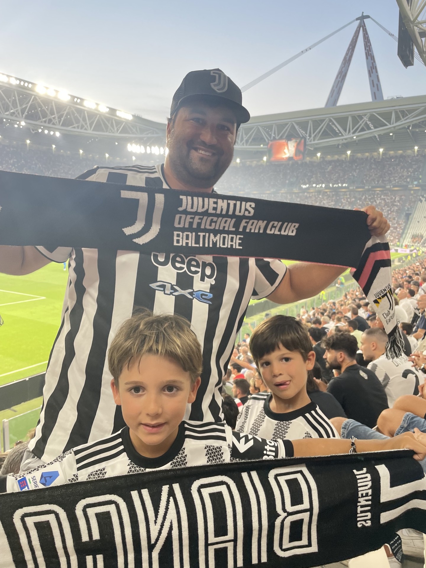 Two children and a man in Juventus football jerseys at a stadium, holding a Juventus scarf, with a stadium background.