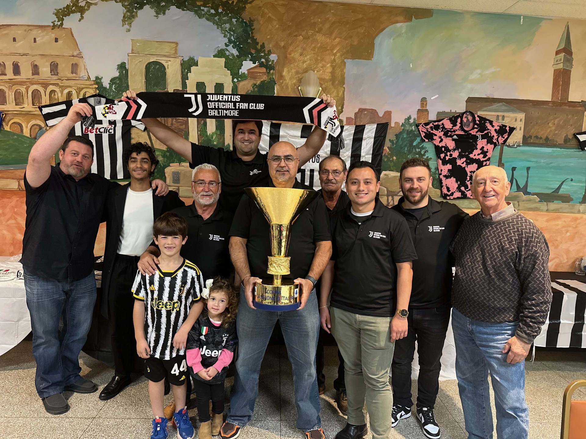 Group of ten people, including children, standing indoors in front of a mural, holding a large trophy, smiling.
