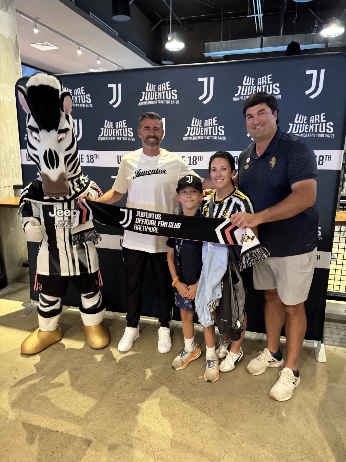 Group of four people, a mascot, and a backdrop with Juventus logos, holding a large check, inside a store or event space.