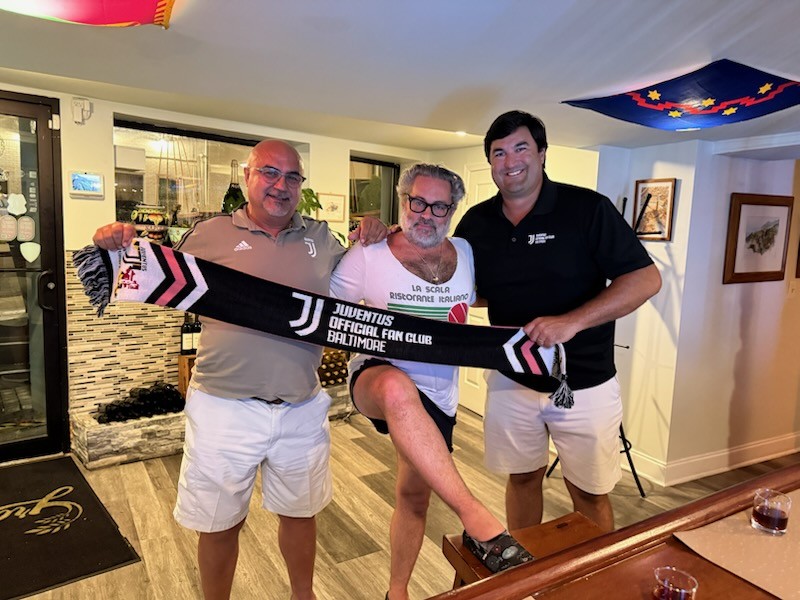 Three men standing indoors, holding a Juventus football club scarf, smiling at the camera.