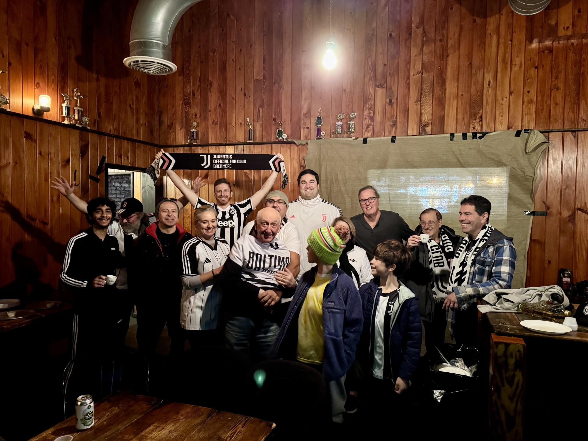 Group of people gathered indoors, some holding a soccer ball, in front of a wooden wall with a projector screen.