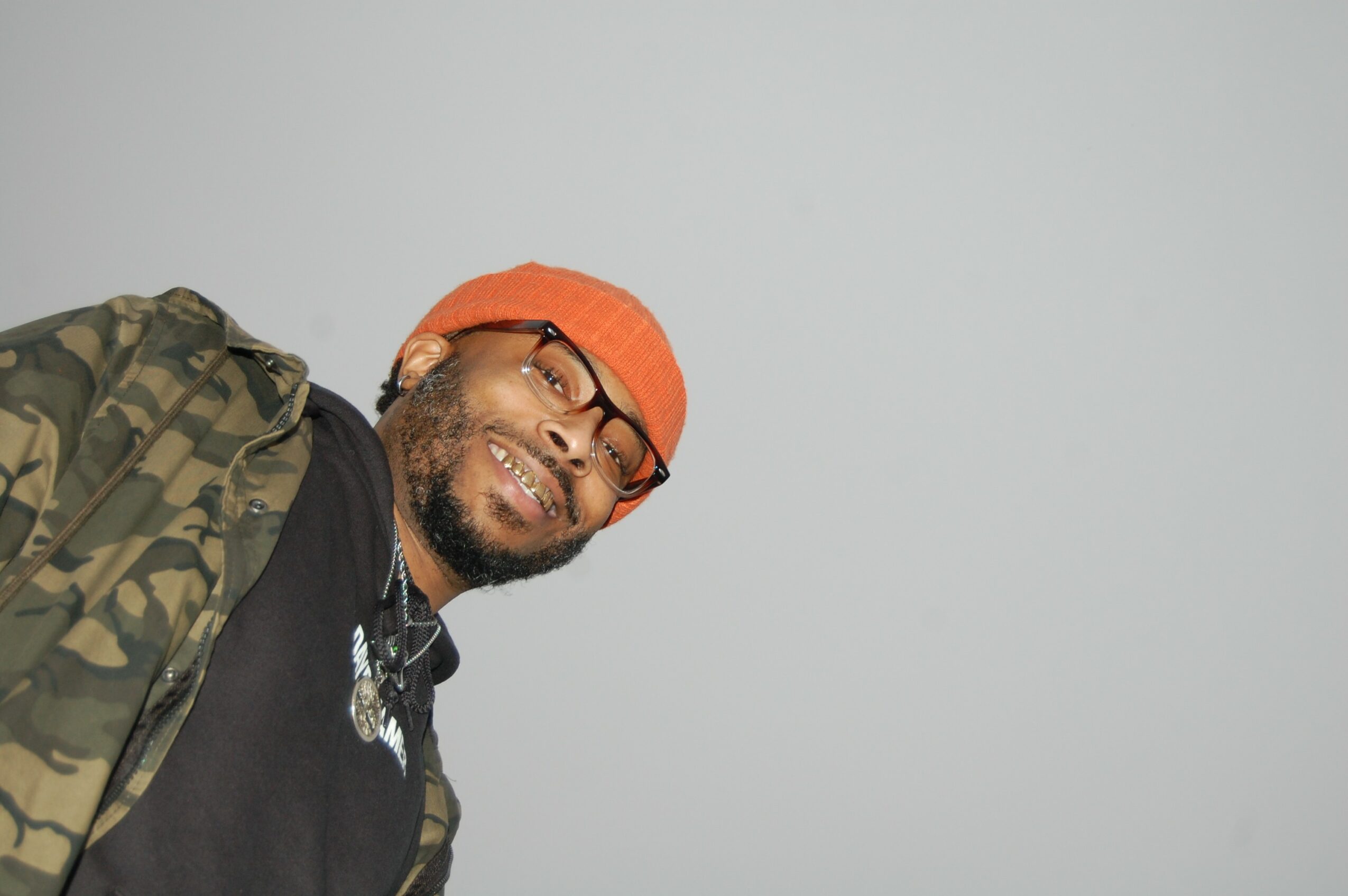 Person wearing glasses, orange beanie, and camouflage jacket smiling, with a plain light gray background.
