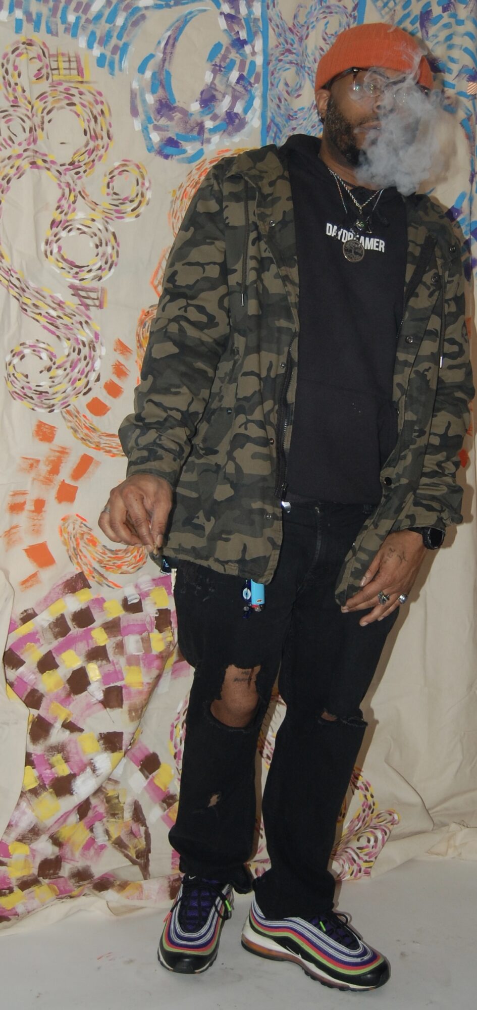 Man with gray beard wearing orange cap, camouflage jacket, black pants, and colorful sneakers standing in front of patterned background.