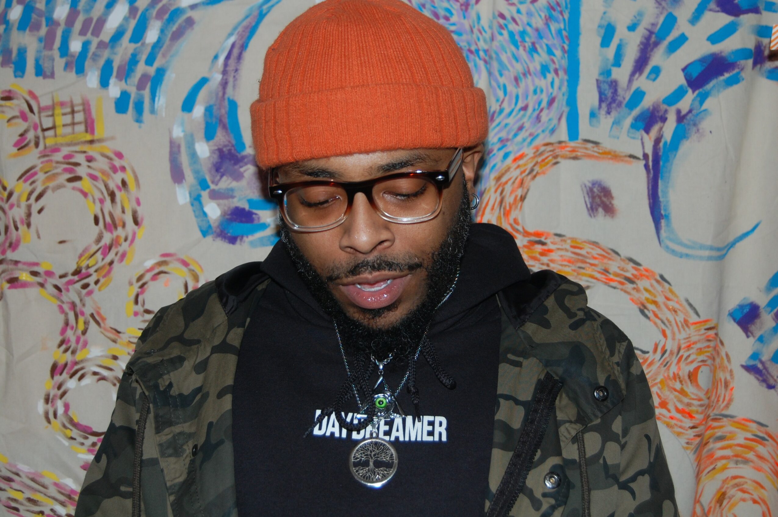Person wearing glasses, orange beanie, black hoodie, and camouflage jacket, standing in front of colorful abstract background.