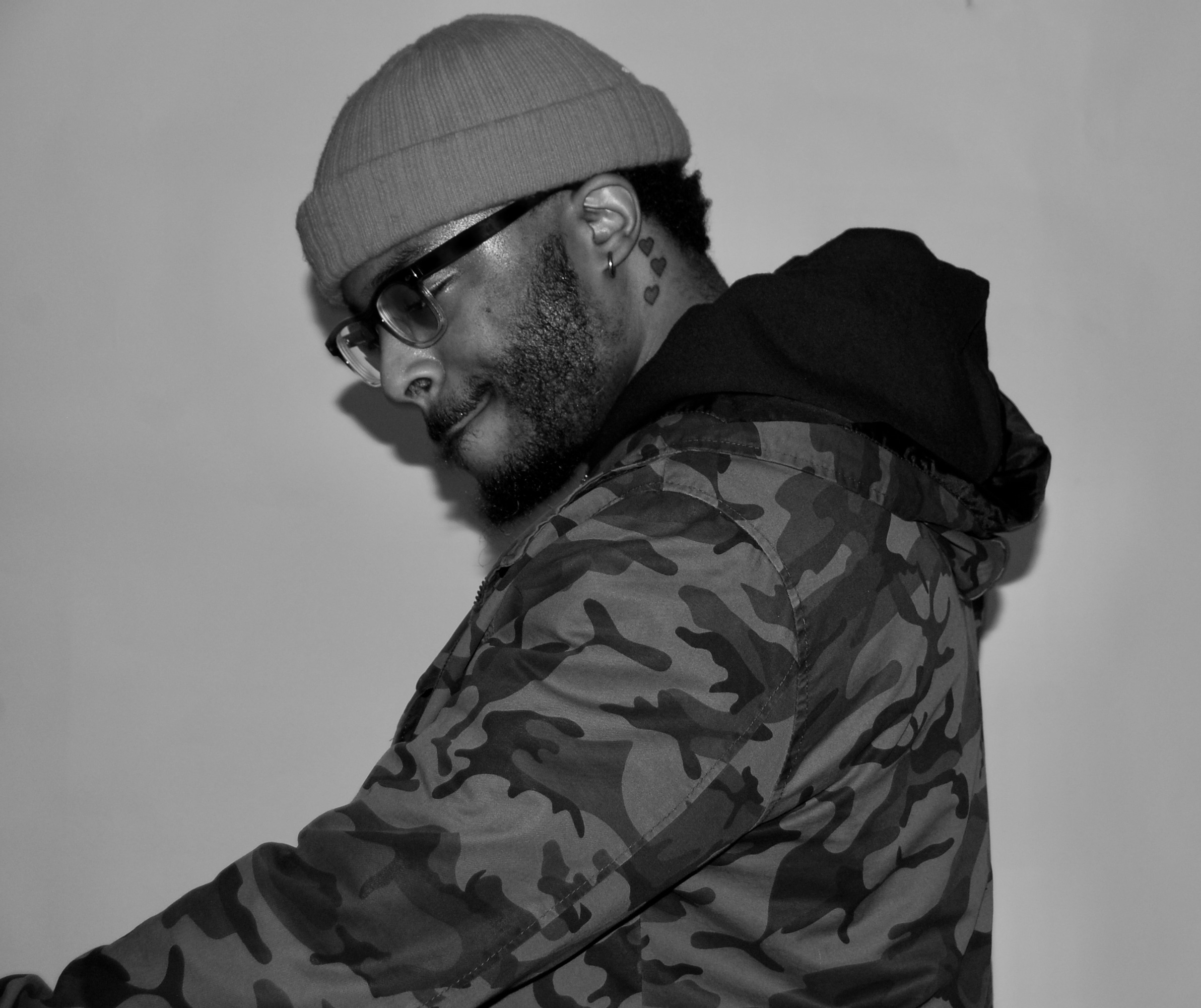 Man wearing glasses, beanie, and camouflage jacket, looking downward, in black and white.