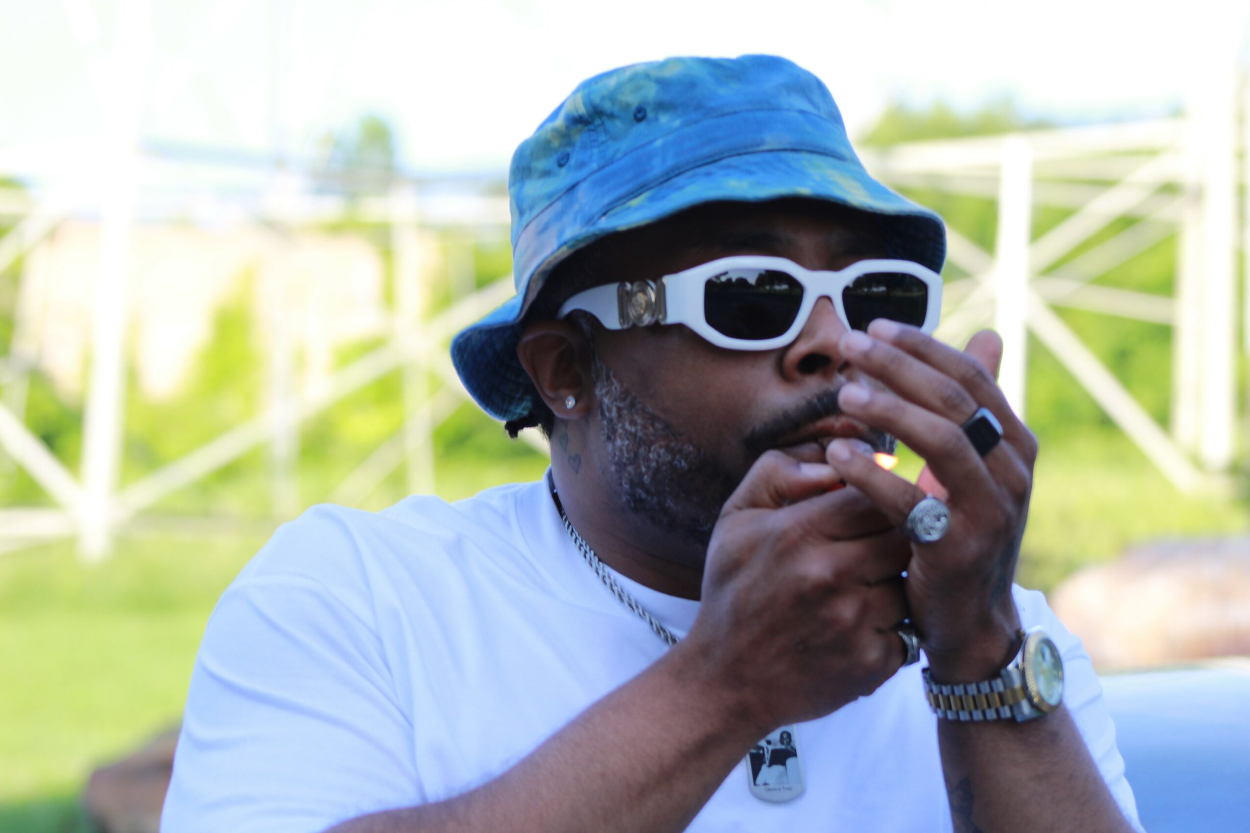Person wearing a blue hat, white sunglasses, and a white shirt, holding a small object near their mouth outdoors.