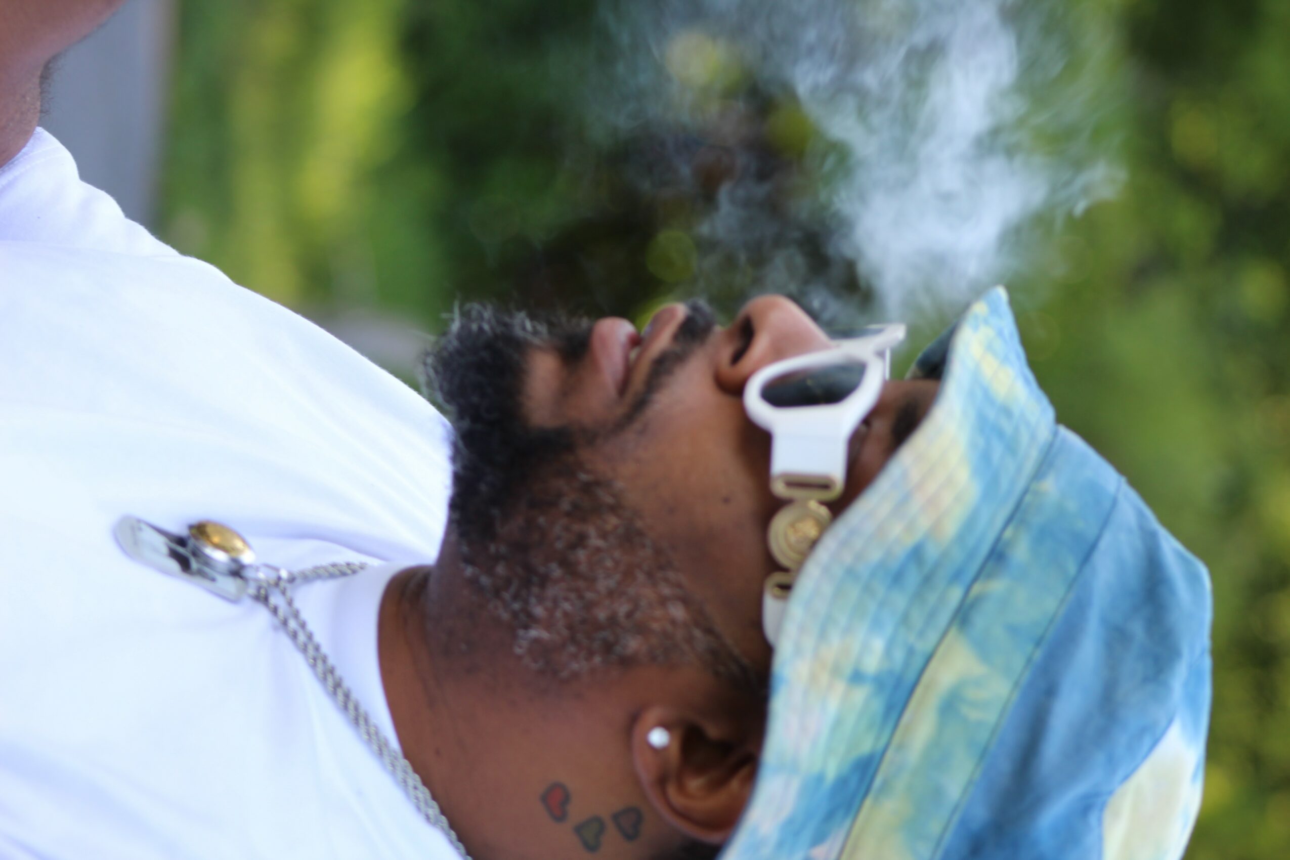 Person lying outdoors with a blue cloth covering their face, smoke rising, and greenery in the background.