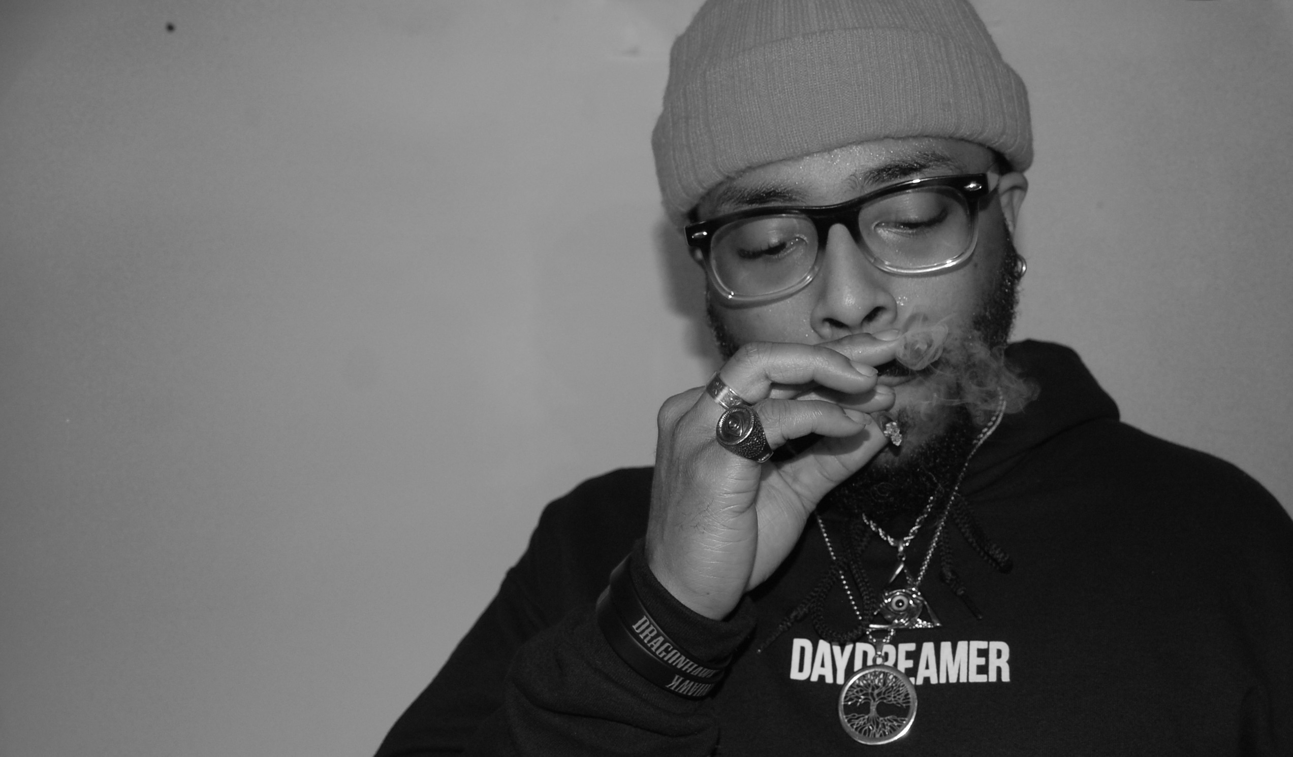 Person wearing glasses, beanie, and hoodie, holding a cigarette near face, with eyes closed, in black and white.