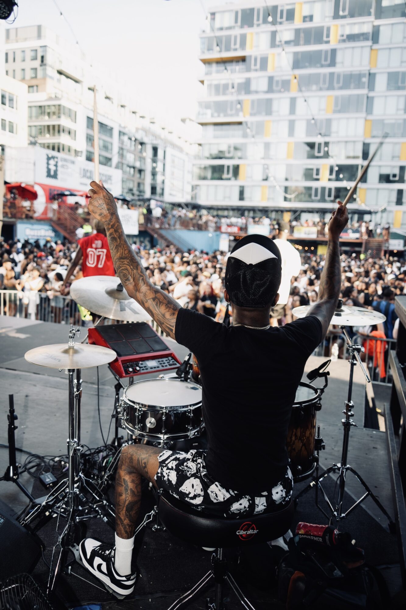 Person playing drums on stage with an audience and city buildings in background, arms raised, wearing a black shirt and white cap.