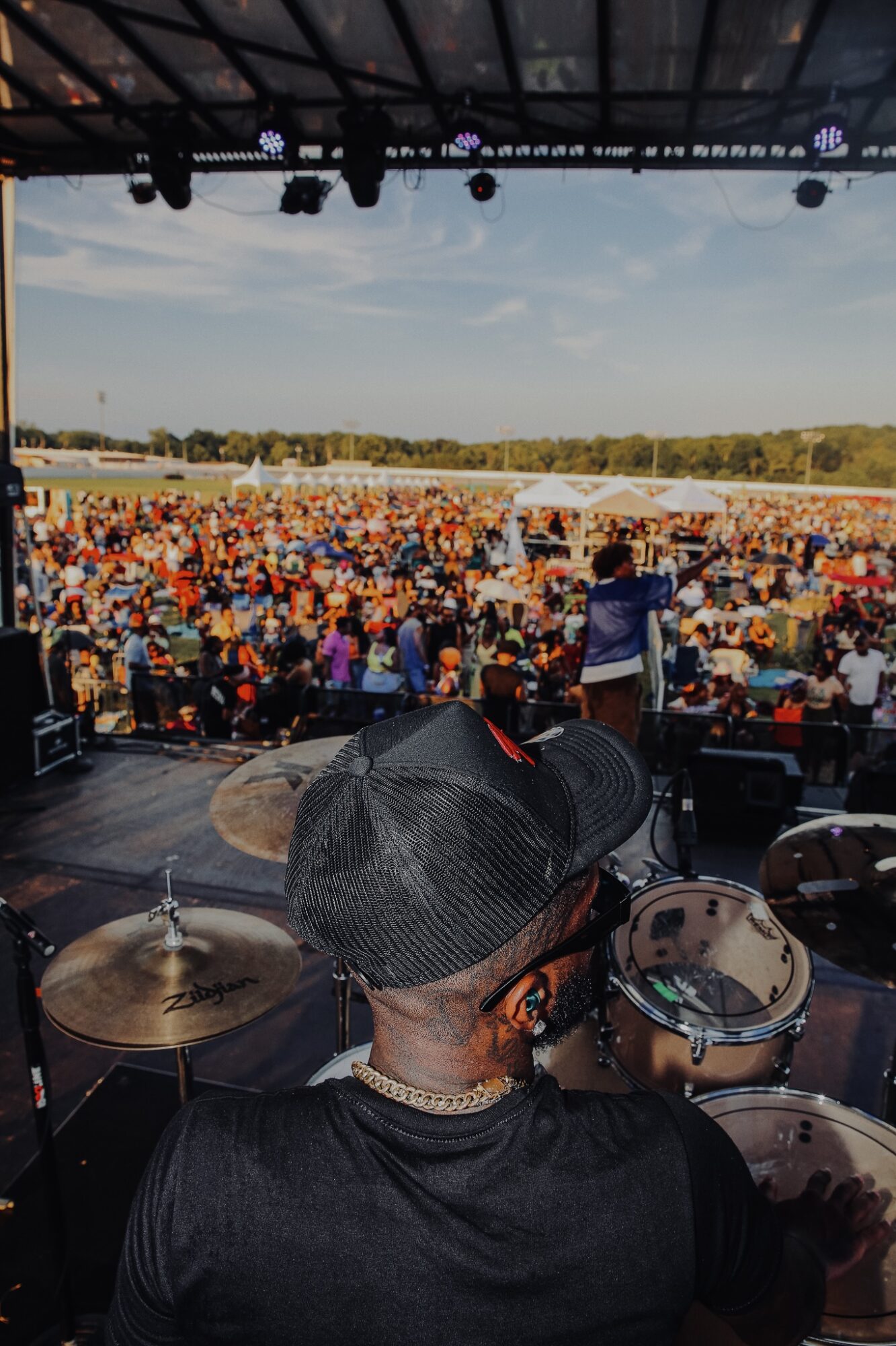 View from stage showing a large outdoor crowd, stage equipment, and a person playing drums, with a landscape and sky in the background.