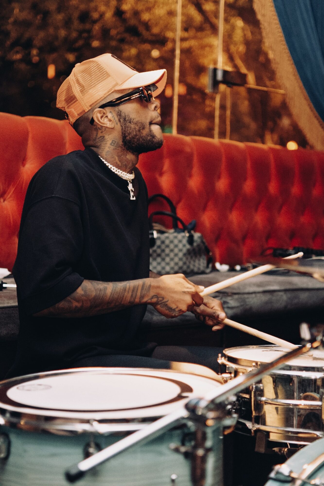 Man with sunglasses, cap, and tattoos playing drums at a venue with red seating and warm lighting.
