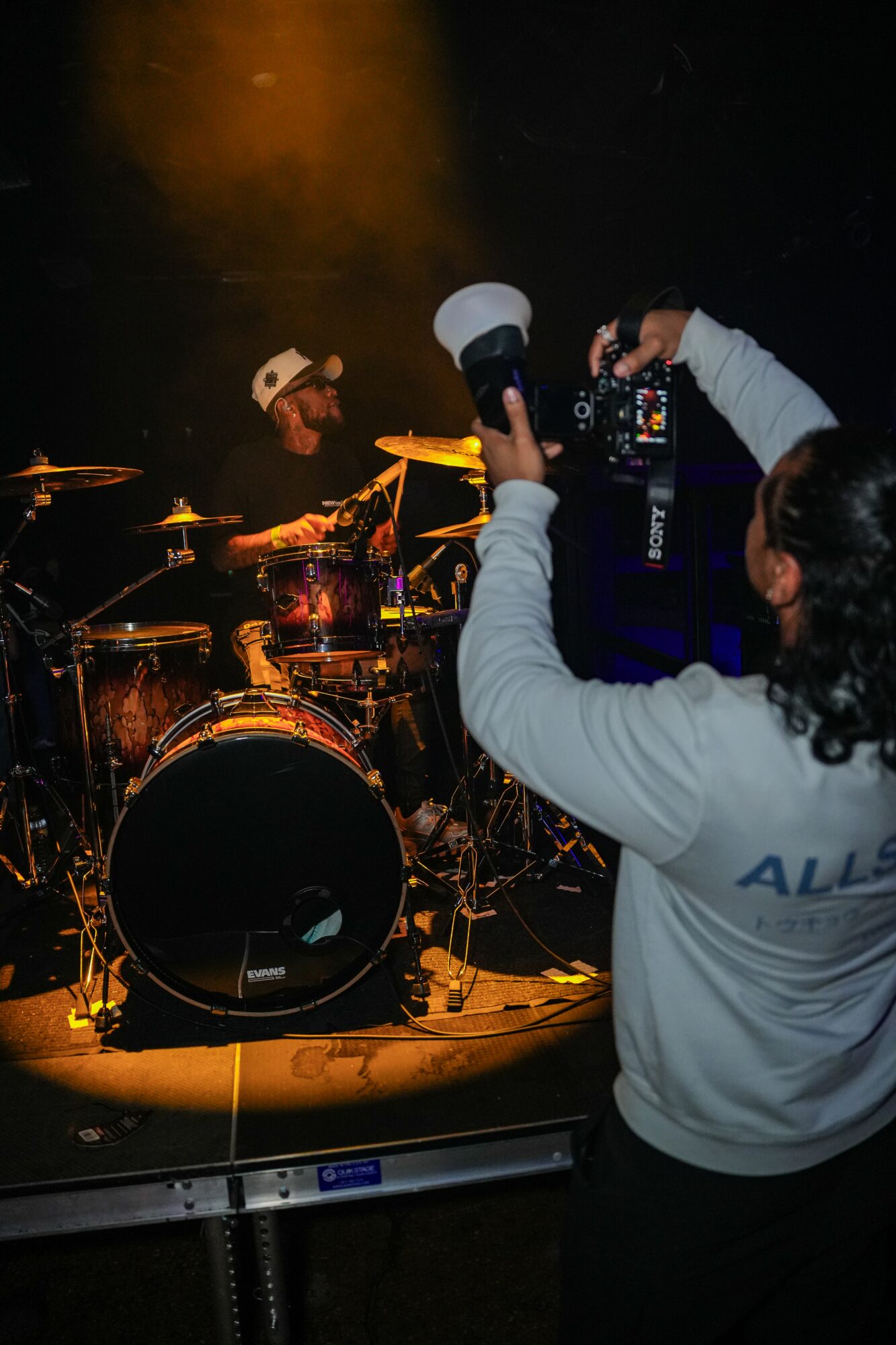 Person taking a photo of a drummer on stage with drums and dark background, wearing a white hoodie, holding a camera, with stage lighting.