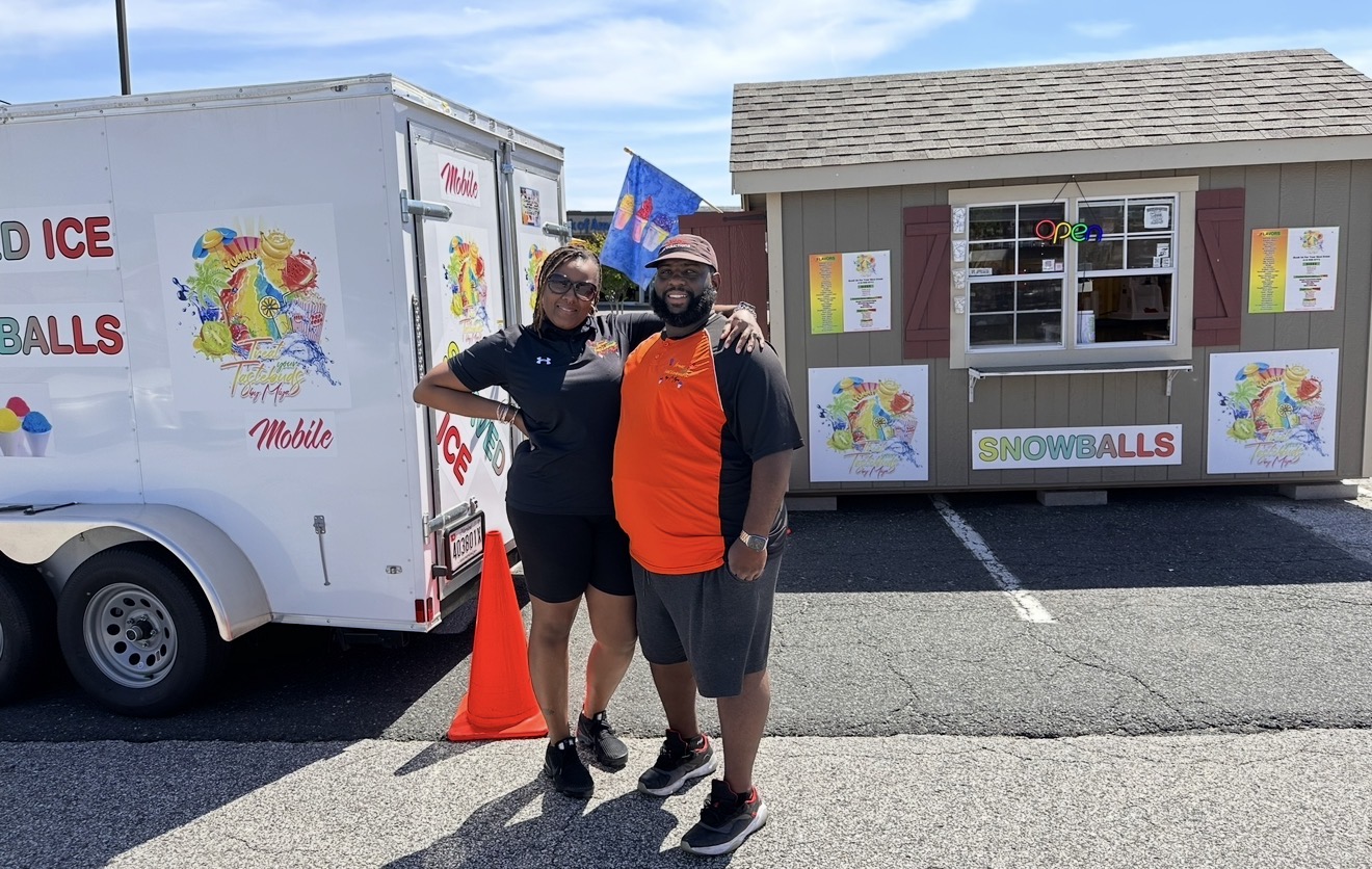Two people standing in front of a snowball stand and a trailer with snowball signs, outdoors on a sunny day.