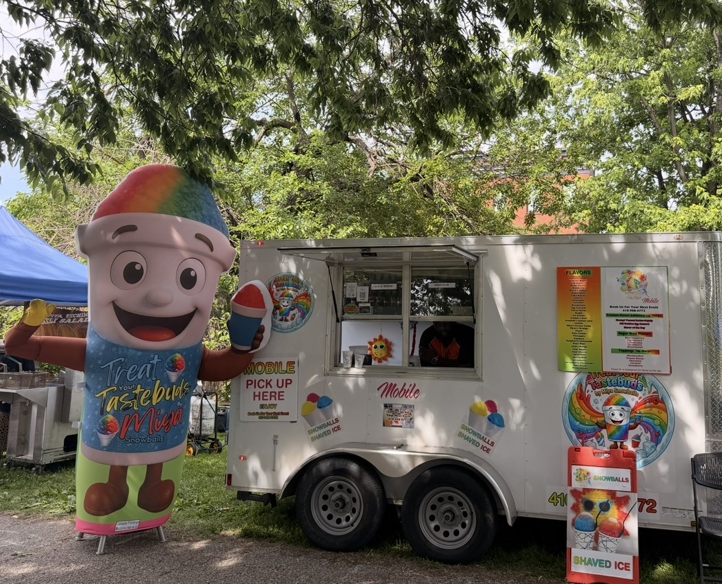 Ice cream truck with colorful mascot outside on a sunny day, surrounded by trees and a blue tent.