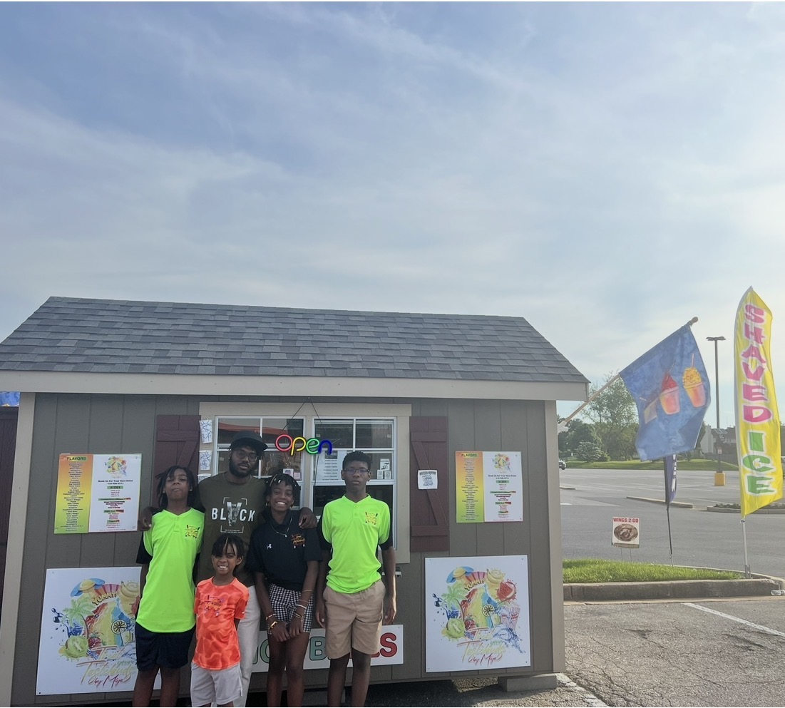 Group of four people, including children, standing outside a small building with colorful posters and flags, under a blue sky.