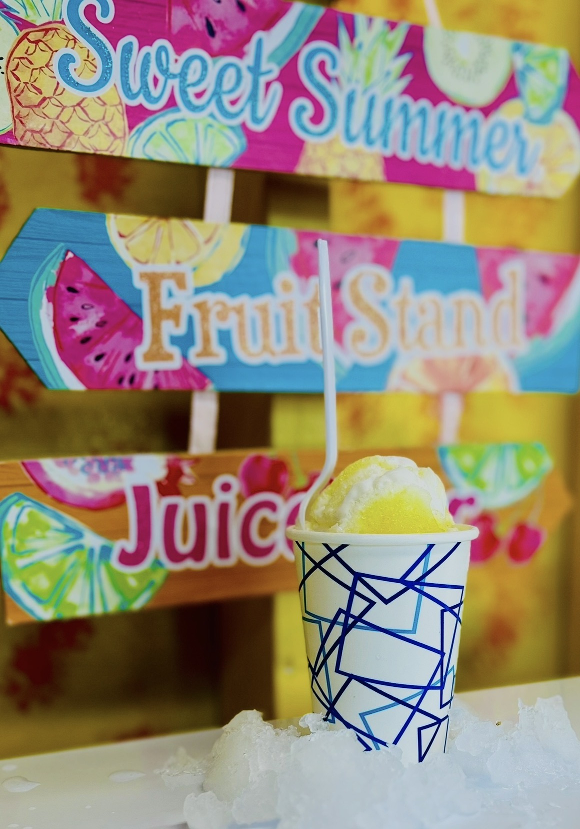 Cup with geometric blue pattern and a scoop of yellow ice cream, in front of colorful signs reading 'Sweet Summer,' 'Fruits,' and 'Juice.'