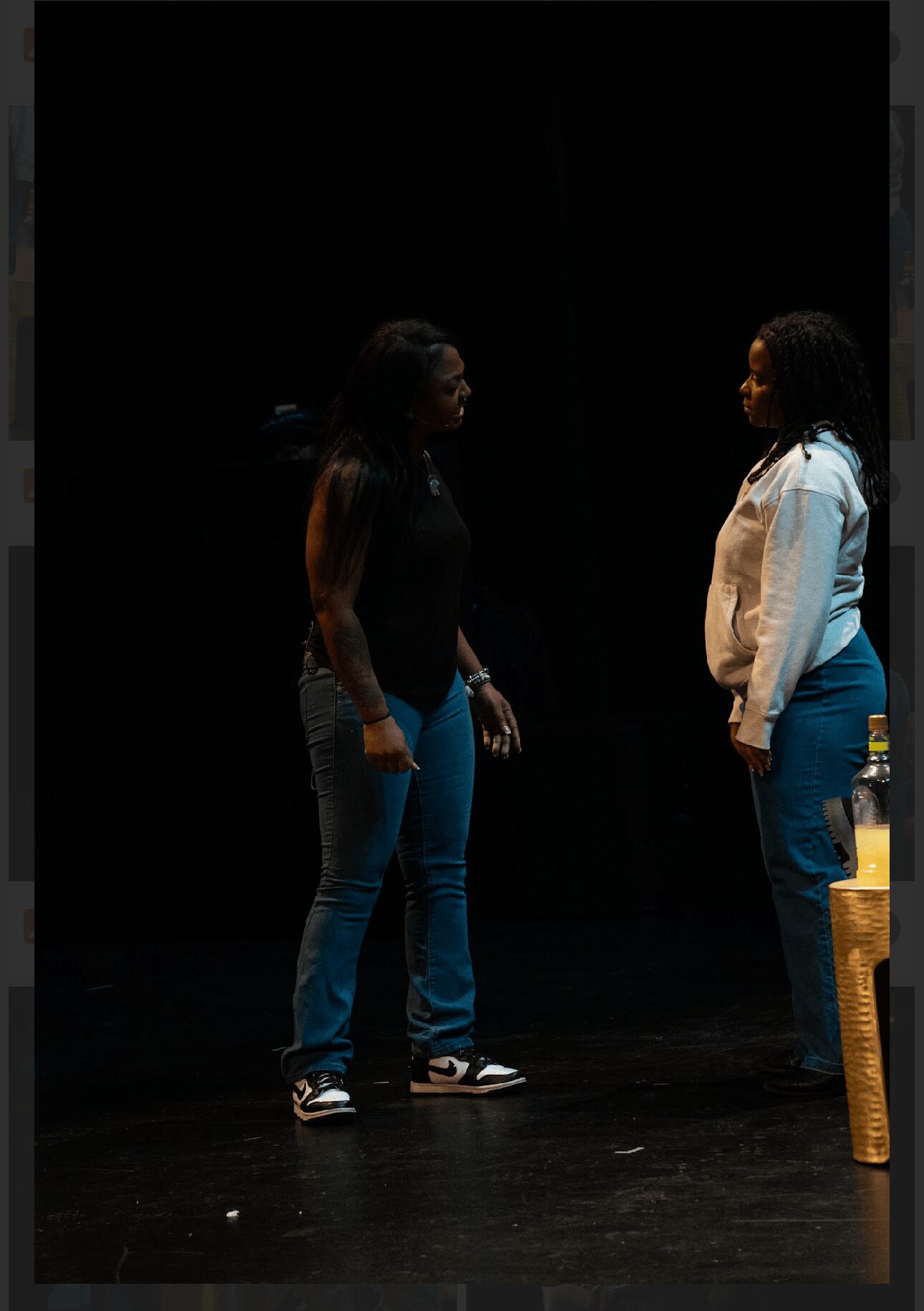Two women stand facing each other on a dark stage, engaged in conversation, with a yellow object nearby.