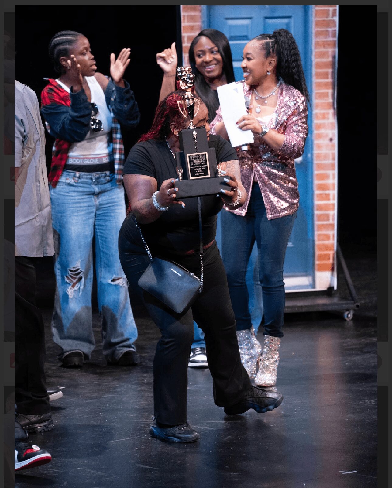 Group of four women celebrating, one holding a trophy, others clapping and smiling, in an indoor setting with a brick wall and blue door.
