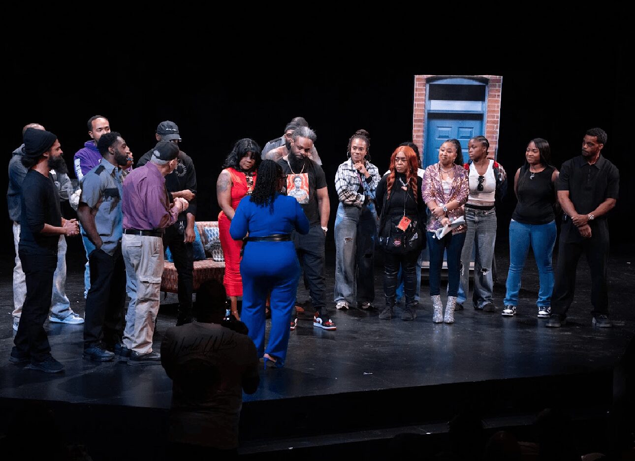 Group of people standing in a semi-circle on a stage, with a person in blue speaking to them, a brick wall and window in the background.
