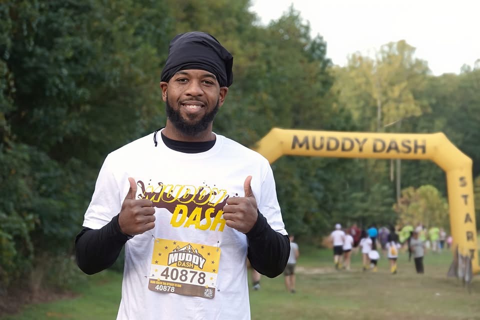 Smiling man in white shirt and black head covering giving thumbs up at muddy dash event, trees in background.