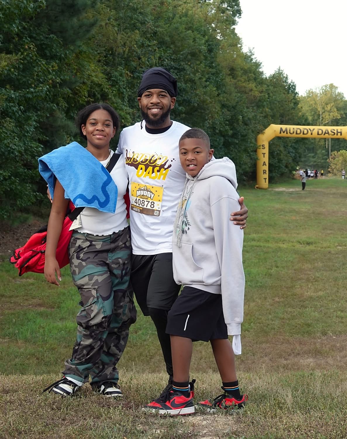 Three people standing outdoors on grass, smiling, with trees and a yellow arch in the background, one person has an arm around each other.