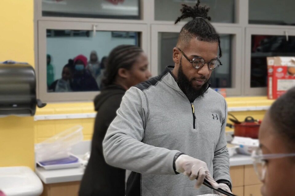 Man with glasses and a beard wearing a gray jacket, working with tools indoors, with people in the background.
