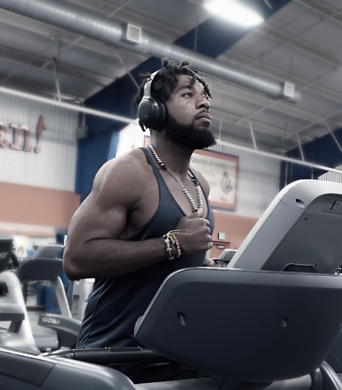 Man running on treadmill in gym, wearing headphones, tank top, and jewelry, with gym equipment and ceiling visible.