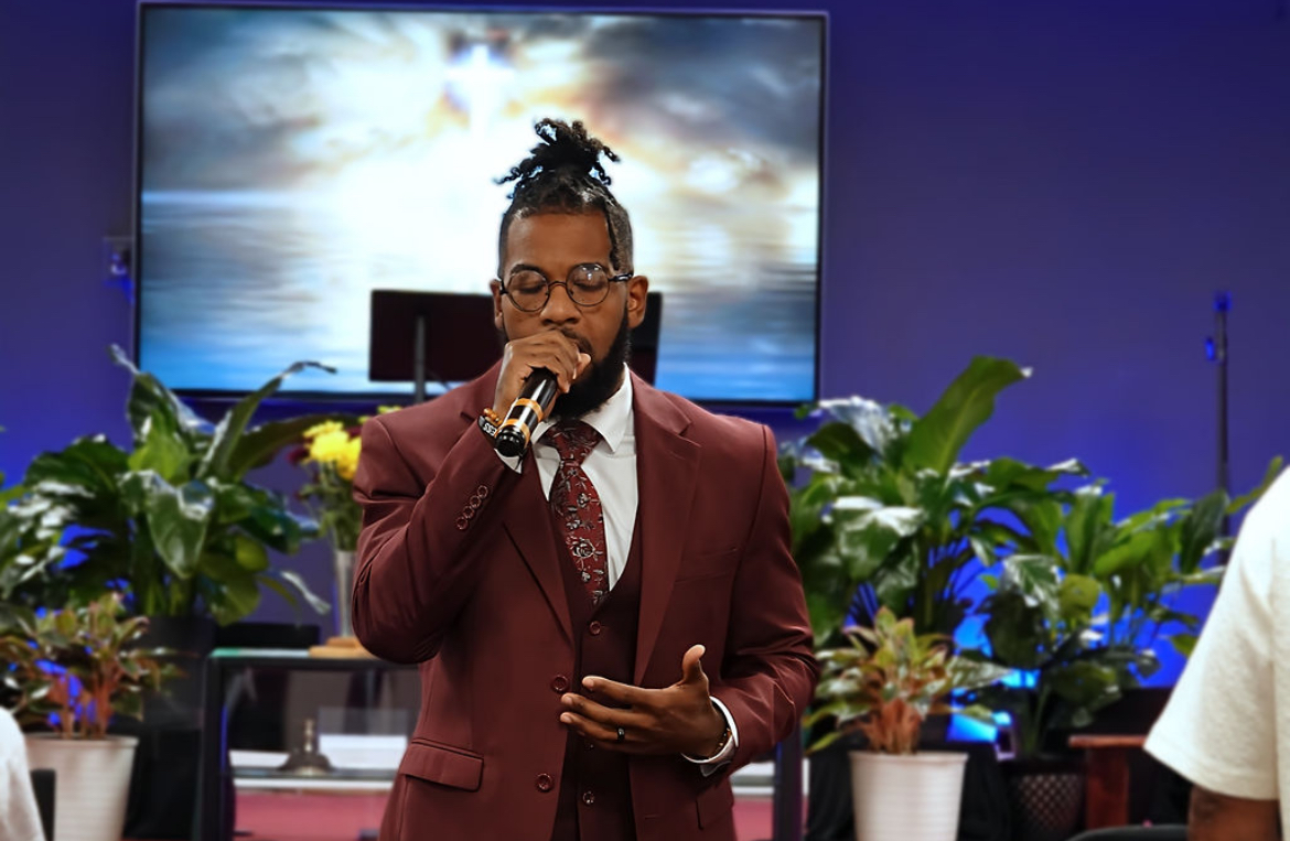 Man with glasses and styled hair singing into a microphone in a room with plants and a large screen behind him.