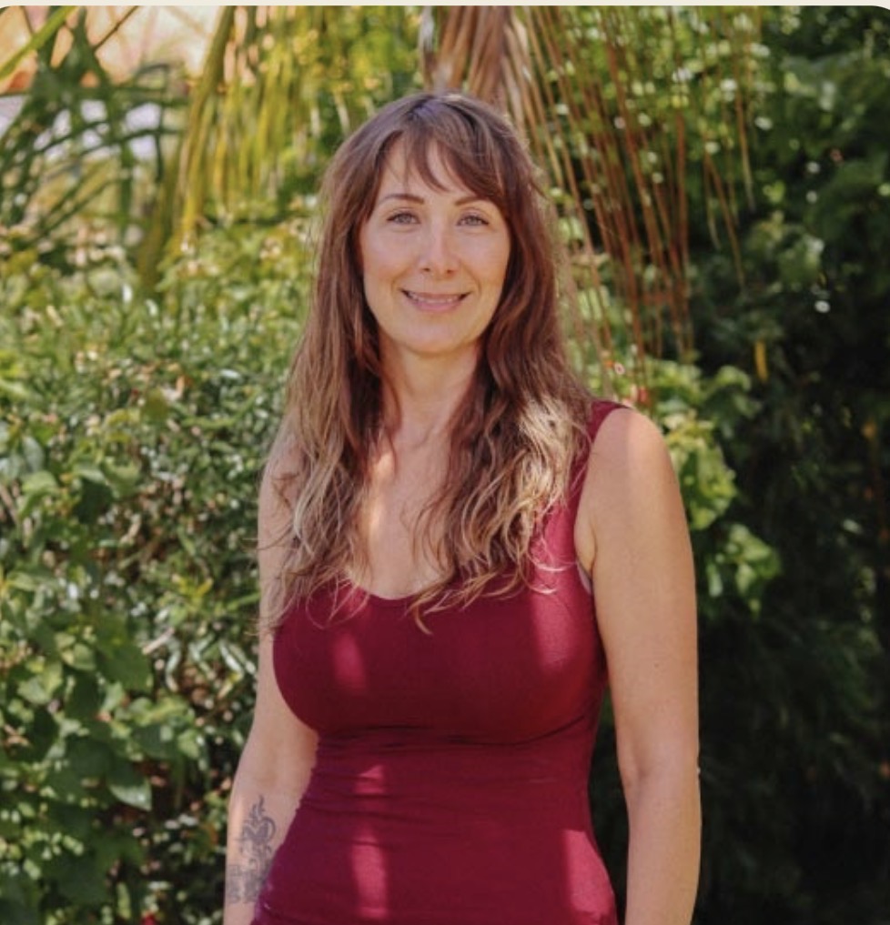 Woman with long wavy hair wearing a sleeveless red top outdoors, smiling, surrounded by green foliage.