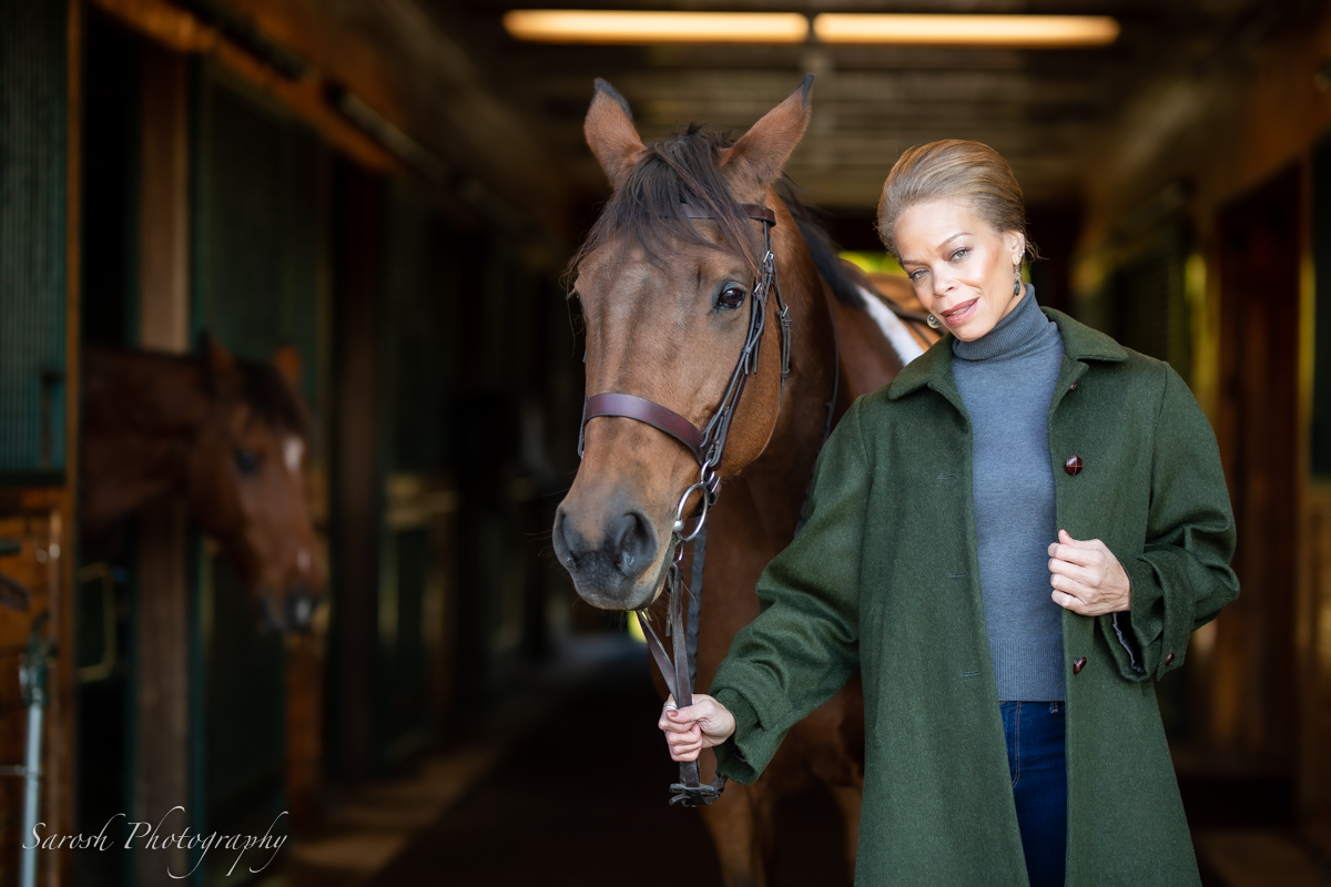 Woman holding a horse's lead in a stable, smiling, with another horse in the background.