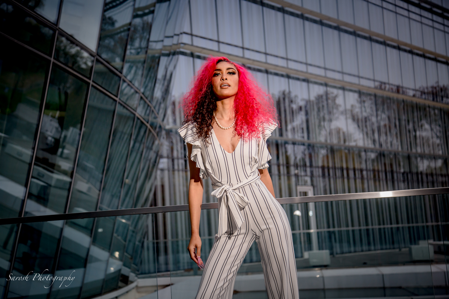 Young woman with bright pink and black curly hair stands outside a modern glass building, wearing a striped jumpsuit.