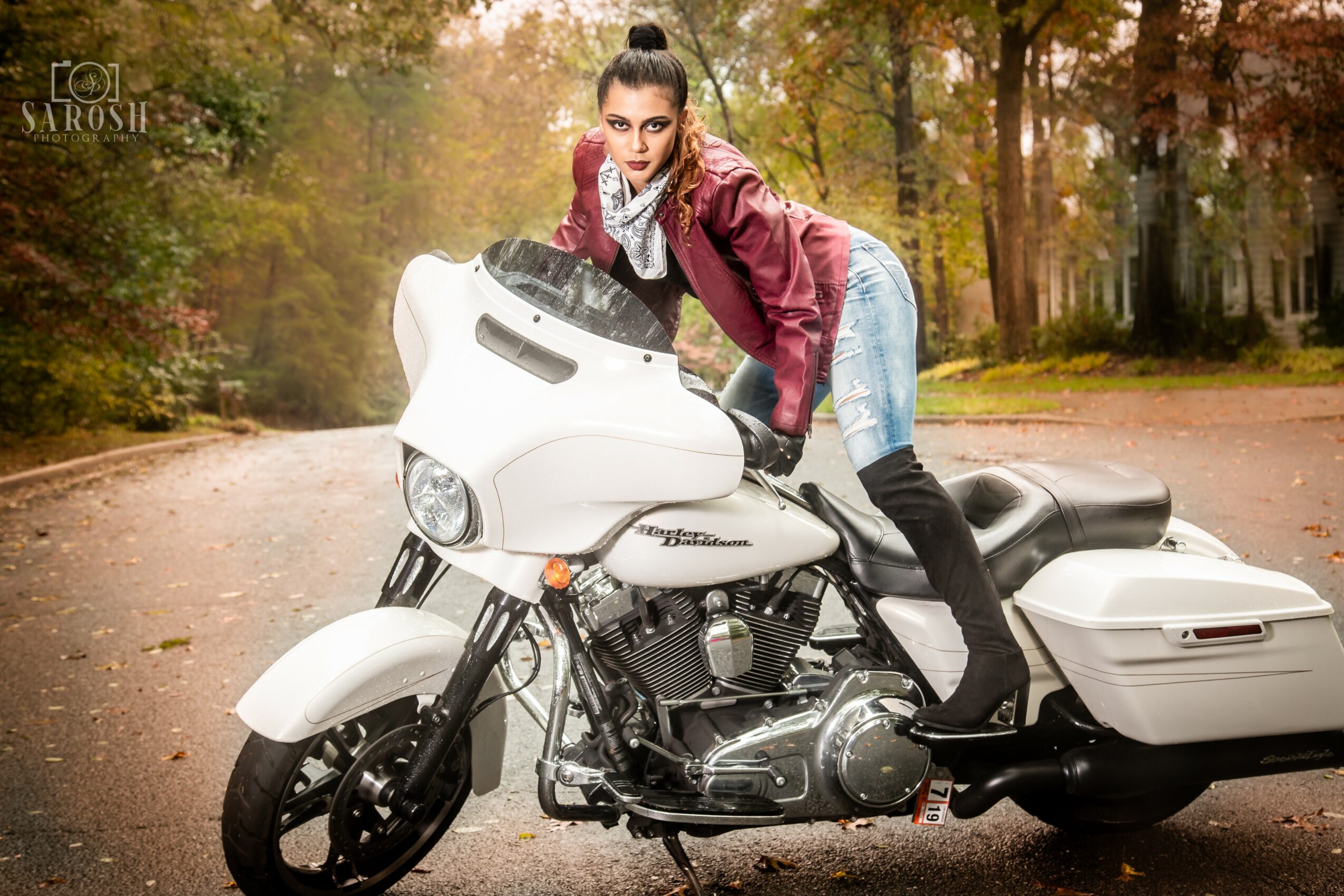 Woman in a pink jacket and jeans leaning on a white motorcycle on a tree-lined road during autumn.