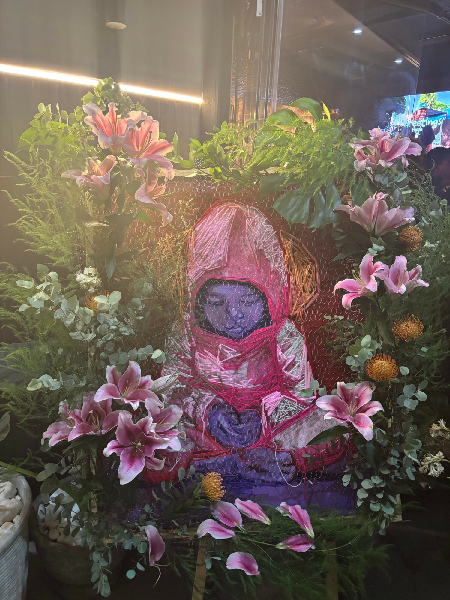A floral arrangement with pink lilies, green foliage, and a central decorative element, possibly a face or mask, surrounded by flowers.